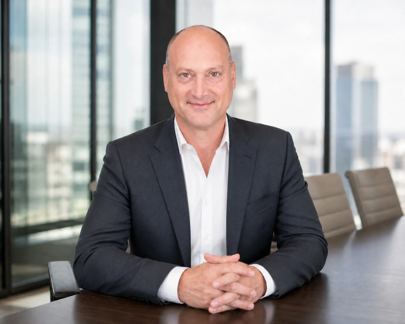 Peter DeCaprio seated at a boardroom conference table with hands clasped, city towers visible through the windows behind him