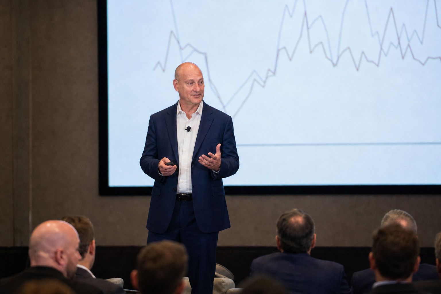 Peter DeCaprio speaking on stage to a seated audience in front of a large screen showing a market line chart